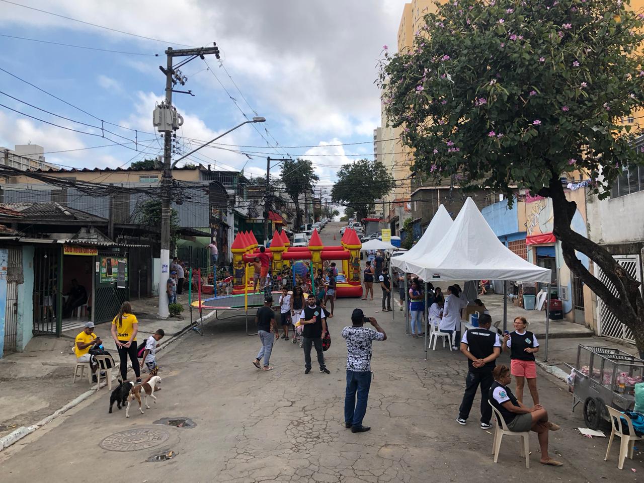 Imagem de cima do palco onde mostra toda a rua do evento com os brinquedos, tenda, carrinho de pipoca e várias pessoas  